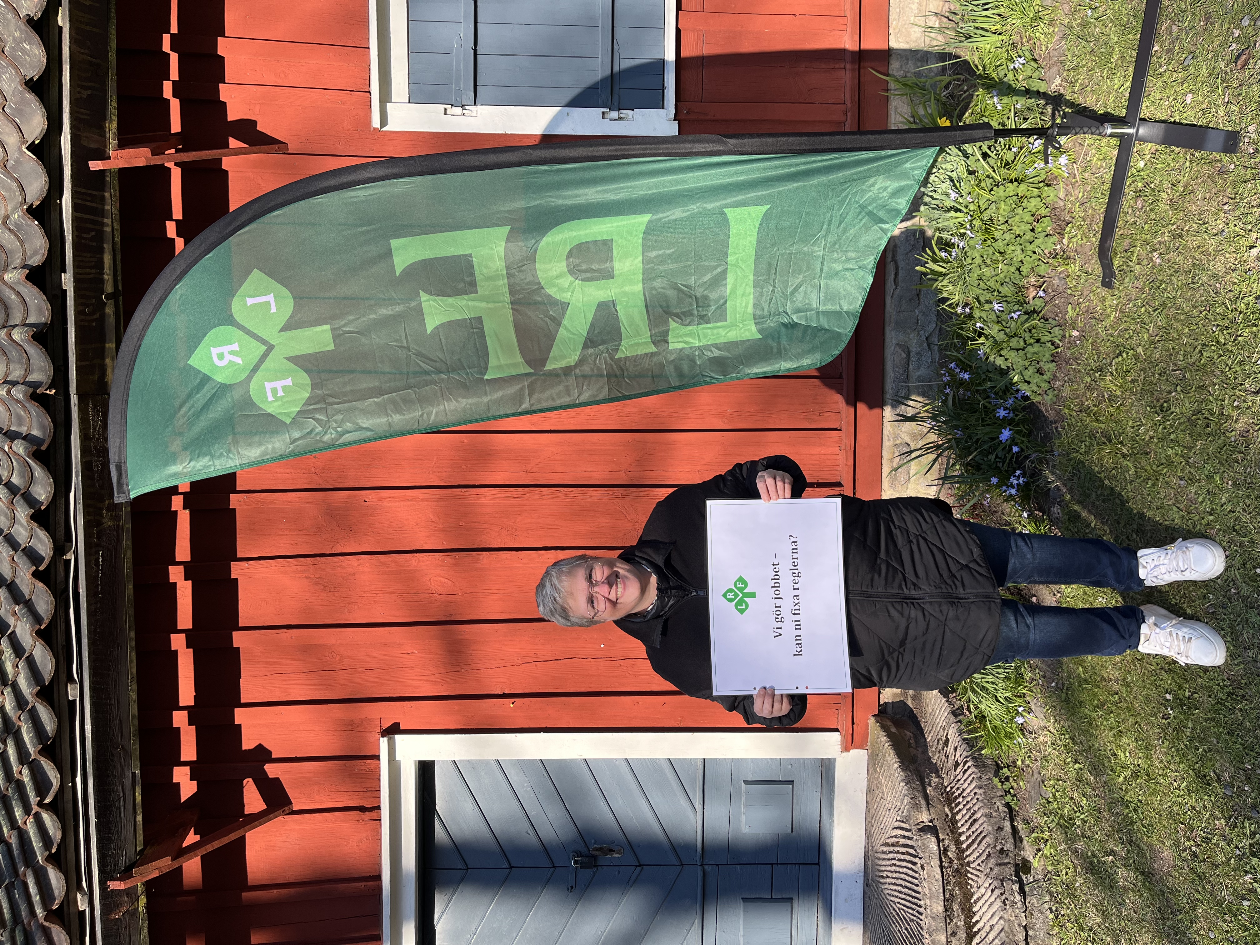 Person står mot en röd husvägg och håller i en skylt med texten "Vi gör jobbet - kan ni fixa reglerna?"